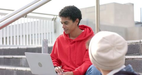 Students collaborating on laptop while sitting on urban campus steps, red hoodie focus