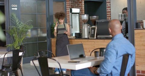 Barista talking on phone while checking tablet at modern espresso counter with laptop customer