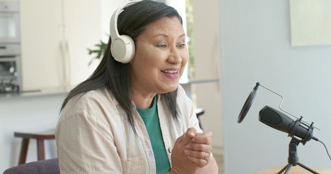 Mature woman hosting home podcast smiling, wearing headphones, speaking into microphone