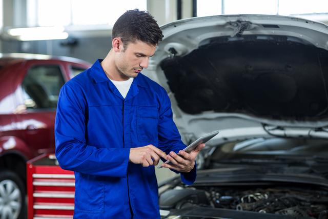 Young male mechanic in blue uniform uses a digital tablet while standing next to a car with its hood open in a repair garage. This image can be used for topics related to automotive repair, technology integration in mechanics, or professional vehicle maintenance services.