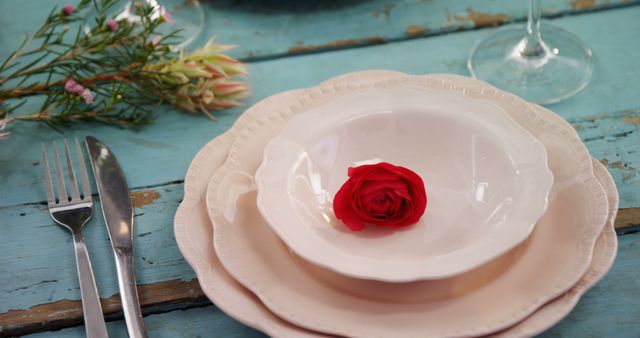 A vibrant red rose rests in the center of an elegant white plate, complemented by silverware and a wine glass on a rustic blue wooden table, with copy space. The setting suggests a romantic meal or a special occasion awaiting the arrival of guests.