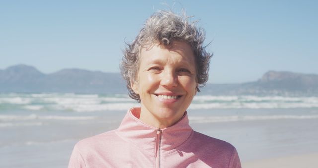 Mature woman with curly gray hair smiling at camera on sunny beach. Ideal for concepts related to happiness, serenity, retirement, and a healthy lifestyle. Can be used in ads promoting vacations, wellness, fitness, and senior activities.