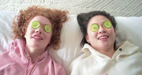 Two friends relaxing with cucumber eye slices on bed in cozy hoodies for self care