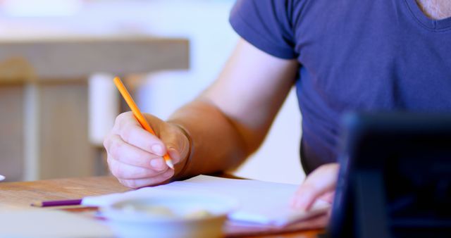 Person holding a pencil, writing on paper at a desk. The scene is likely engaged in studying, work, or completing assignments. Ideal for educational blogs, productivity articles, and office setup highlights.