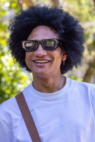 Sunlit african man smiling in bold sunglasses and white tee, urban park portrait with afro