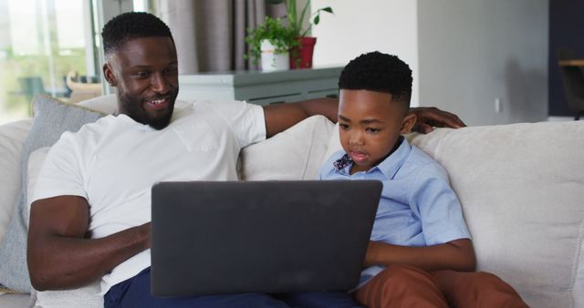 Father and son sitting on couch, using laptop together. Ideal for illustrating family bonding, parenthood, educational websites, technology usage in the family, and home leisure activities.
