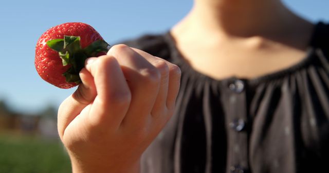 Person is holding a ripe strawberry outdoors in sunny weather. This image is perfect for advertising healthy eating habits, summery recipes, lifestyle blogs, and nutritional articles. It can also be used for promotion of organic or local farming products and appealing to nature enthusiasts.