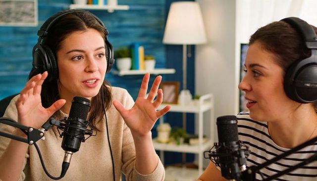 Two female podcasters recording conversation in cozy home studio with headphones and mics