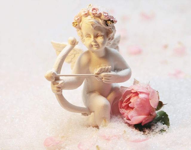 Cute Cupid figurine holding bow and arrow sits on winter snow surrounded by petals and rose. Perfect for Valentine's Day, wedding decor, holiday greeting cards, and romantic themes.