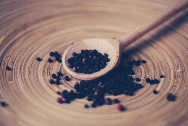 Closeup of black peppercorns spilling from wooden spoon on swirled wooden plate. Ideal for cooking blogs, culinary stock photos, food preparation visuals, natural and rustic advertising, and seasoning packaging designs.