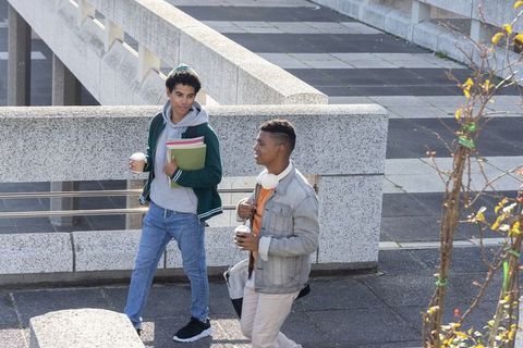 Two diverse college students walking campus bridge carrying notebooks, backpacks, coffee