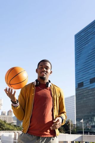African american man spinning basketball in city plaza wearing headphones and mustard bomber jacket
