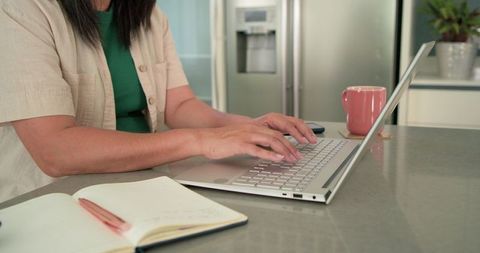 Female remote worker typing on laptop at kitchen counter with notebook and pink mug