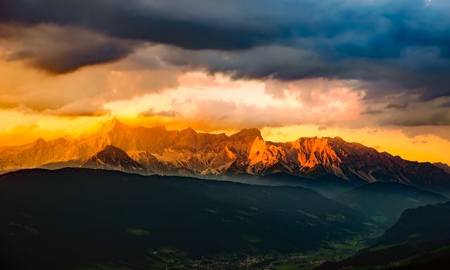 This stunning visual captures the vibrant colors of the setting sun illuminating a majestic mountain range. The dramatic clouds add depth and contrast, making it perfect for promotional material focused on travel, outdoor recreation, or inspirational themes. Ideal for use as a background, poster, or website banner related to nature and adventure.