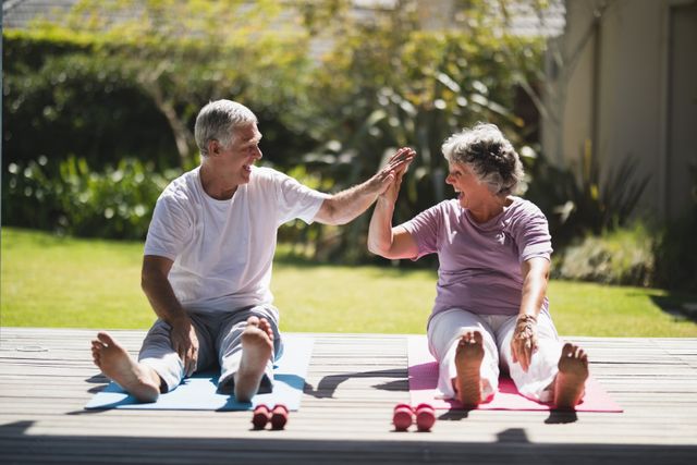 Senior couple sitting on yoga mats on a porch, giving each other a high five while exercising. They are smiling and appear to be enjoying their workout. This image can be used for promoting senior fitness, healthy lifestyles, retirement activities, and wellness programs.