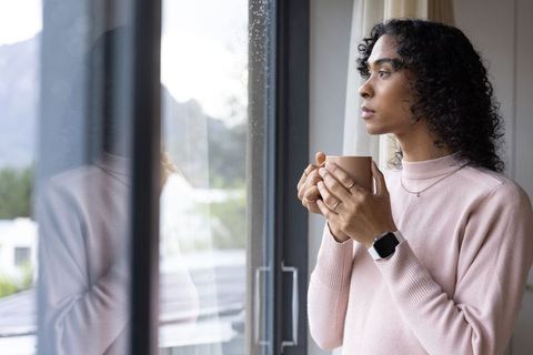 Non-binary person holding mug and gazing through rain-dotted window, wearing pale pink pullover