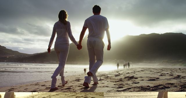 A young Caucasian couple walks hand in hand along a sandy beach at sunset, with copy space. Their relaxed stroll by the sea creates a romantic and serene atmosphere.