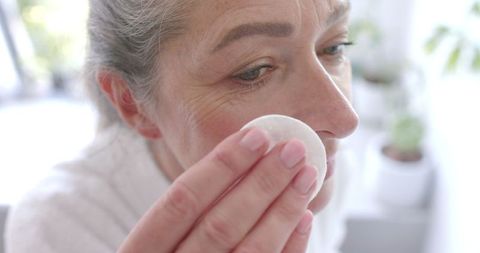 Mature woman cleansing face with cotton pad in domestic setting