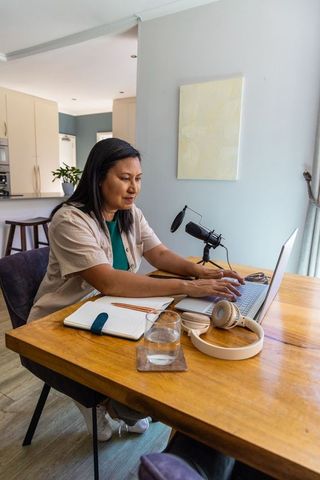 Asian woman working from home with laptop and microphone in open-plan kitchen workspace