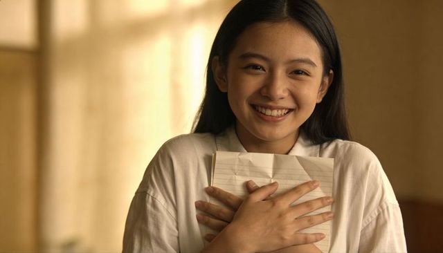Smiling asian teen holding folded lined paper to chest in warm window light, joyful student portrait