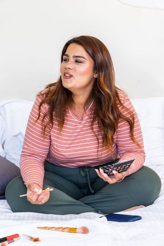 Woman applying makeup on bed during relaxed self-care routine with smartphone