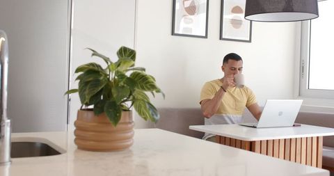 Young man working remotely in sunlit modern kitchen nook drinking coffee