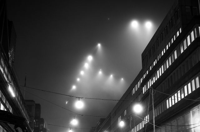 Urban city street at night enveloped in dense fog, illuminated by overhead street lights. The black and white color scheme adds a dramatic and atmospheric effect. Suitable for themes related to urban life, nightlife, atmospheric scenes, and architectural photography.