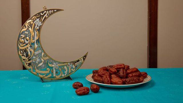 Displaying golden arabic crescent with calligraphy beside plate of dates on turquoise table