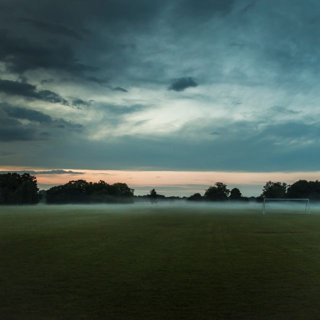 Misty dawn enveloping a serene soccer field with dramatic sky and light creeping through the clouds. Trees and horizon blend in the dawn light, creating a tranquil, atmospheric scene. Suitable for use in projects related to nature, weather, sports, and outdoor activities or conveying calm and serenity in visual designs.