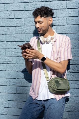 Indian man cradling small brown puppy with headphones and crossbody bag by blue brick wall