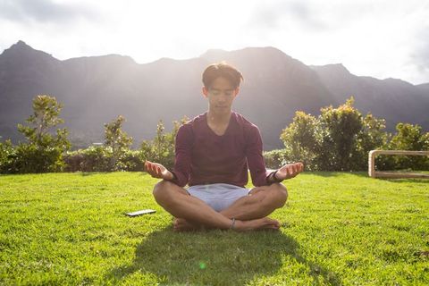 Young asian man meditating outdoors on grass at sunrise with mountains and sunlight