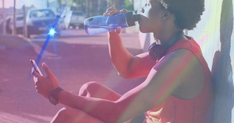 Man drinking water after run on sunlit urban sidewalk with smartphone and headphones