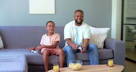 African american father and teenage son playing video games on cozy gray sofa, smiling