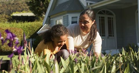 Mother and daughter gardening in sunlit front yard tending purple flowers and learning