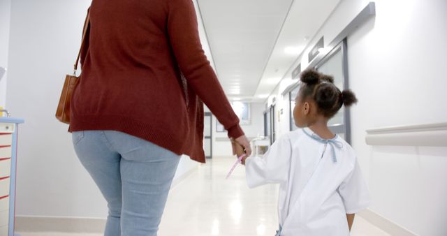African american girl patient and her mother walking holding hands in corridor in hospital. Medicine, healthcare and hospital, unaltered.