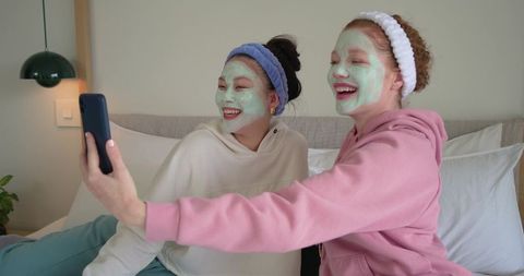 Diverse friends taking skincare selfie wearing green face masks and cozy hoodies on bed
