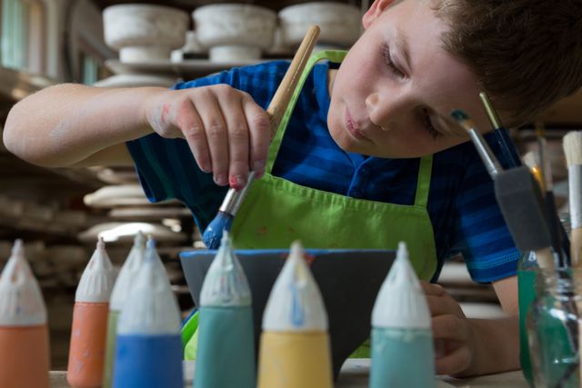 Young boy focused on painting a ceramic bowl in a pottery shop. He is wearing a green apron and using a paintbrush to apply blue paint. Various paint bottles are visible in the foreground. This image is ideal for illustrating children's art classes, creative workshops, and hands-on learning activities.