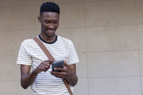Young african man checking smartphone while smiling, wearing crossbody bag by tiled wall