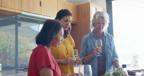 Multicultural women tasting wine and inspecting produce at sunlit modern kitchen island