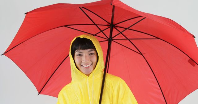 Smiling woman in yellow raincoat holding red umbrella. Ideal for use in content related to rain gear, joyful moods on a rainy day, and vibrant color arrangements. Suitable for marketing rainy weather products, lifestyle blogging, and advertisements focusing on happiness and positivity despite adverse conditions.