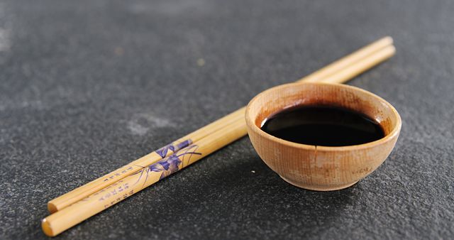 Bamboo chopsticks placed neatly beside small ceramic bowl filled with soy sauce, arranged on a sleek dark slate table. Perfect for illustrating Japanese or Asian culinary themes, traditional dining setups, restaurants, or articles about Asian cuisine. Also suitable for minimalist tableware articles and food presentations.