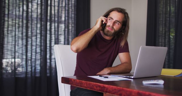 Young bearded man in dark red t-shirt casually talking on phone while working on his laptop in a home office environment. He is sitting at a wooden desk with paperwork spread out, pens and a remote control beside him. Image can be used for concepts related to remote work, freelancing, home office setup, communication, professional life, and balancing work from home.