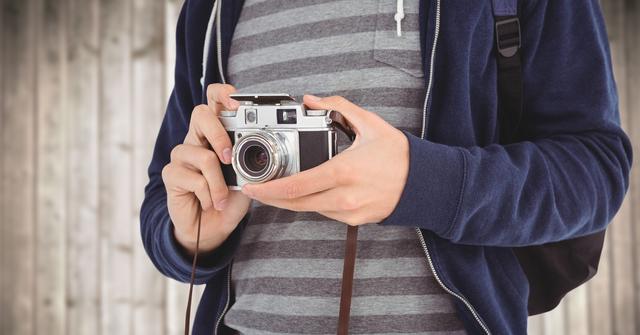 Close-up of man's hands holding vintage camera while standing in casual clothing. Combined with wooden plank backdrop, it evokes creativity and passion for photography. Useful for themes around vintage aesthetics, hobbies, and outdoor activities.