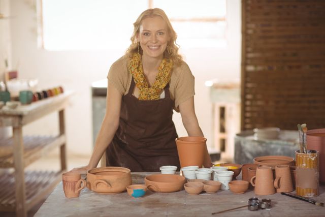 Smiling female potter leaning on worktop surrounded by various handmade pottery pieces in a well-lit workshop. Ideal for use in articles or advertisements related to pottery making, artisan crafts, creative studios, small business promotion, and artistic hobbies.