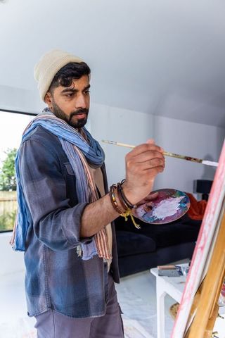 Asian man painting on stretched canvas in bright home studio holding palette and brush