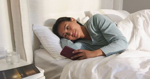 Asian woman relaxing in bed using smartphone in sunlit minimalist bedroom for calm lifestyle