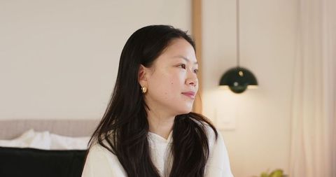 Serene asian woman gazing from cozy minimal bedroom wearing white hoodie and gold hoops