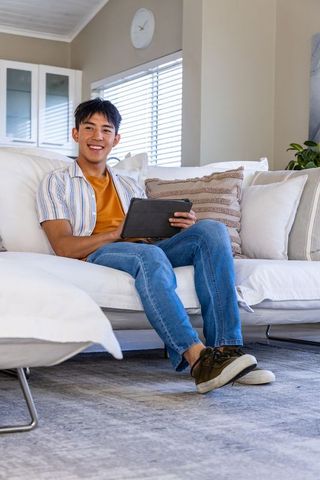 Asian man relaxing on white sofa using tablet in sunlit modern living room