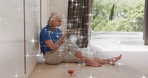 Senior woman sitting on floor using tablet barefoot with tea near balcony and sunlight