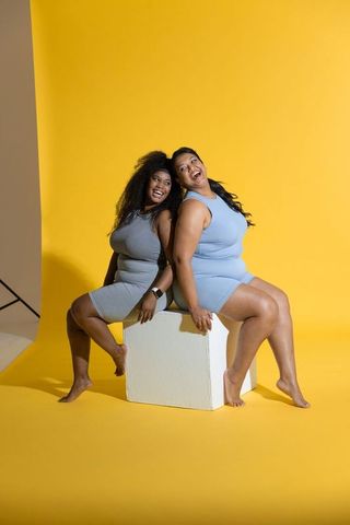 Laughing plus-size women in activewear sitting back-to-back on cube on yellow background